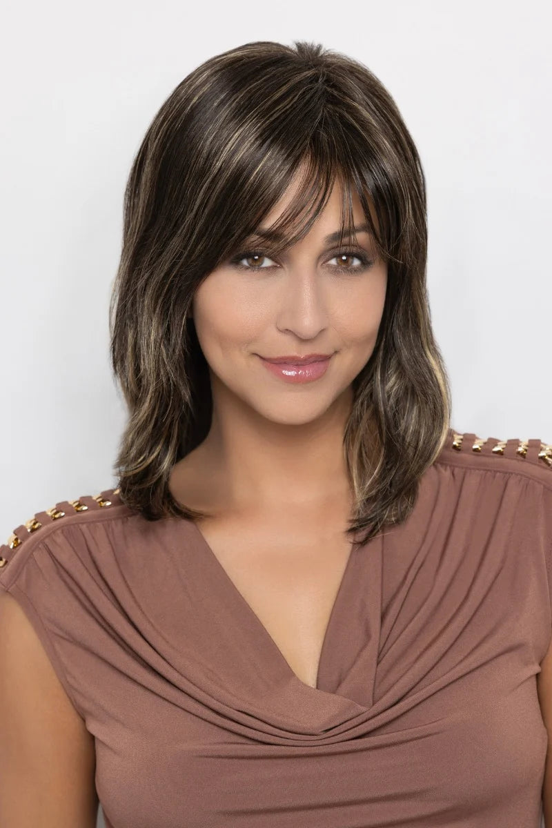 A woman with a shoulder-length brown wig with lighter brown highlights and straight-cut bangs, wearing a draped brown top against a light gray background.