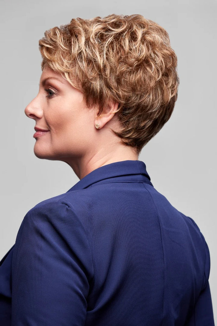 A woman shown in profile wearing a short, layered, curly wig in warm blonde and light brown tones, paired with a navy blue collared top. The background is a solid light gray studio backdrop.