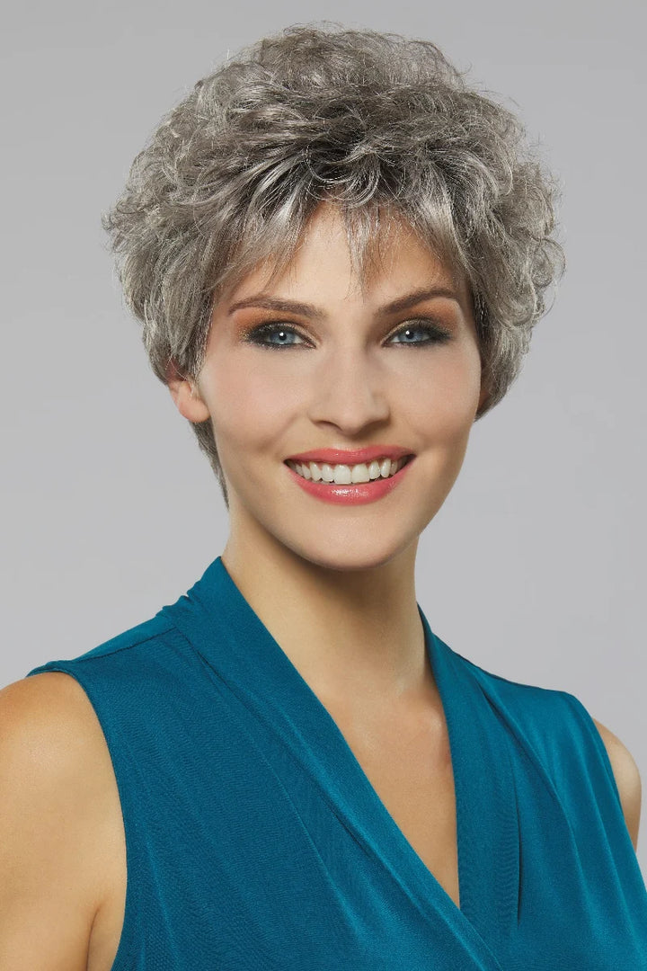 A smiling woman with short, curly gray hair wearing a sleeveless teal top against a plain light gray studio background.