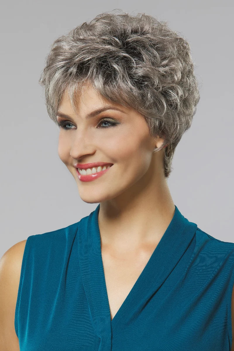 A woman wearing a short, curly gray wig and a teal sleeveless top, shown against a plain light gray studio background.