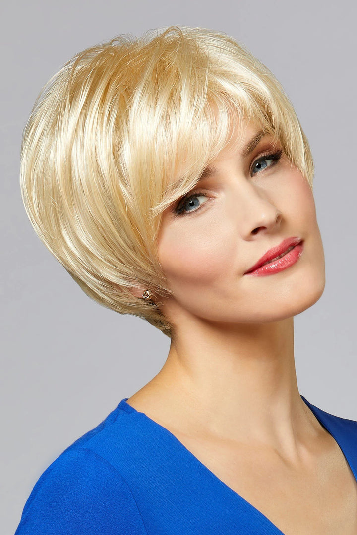A woman wearing a short, layered blonde wig with side-swept bangs, shown against a plain light gray studio background.