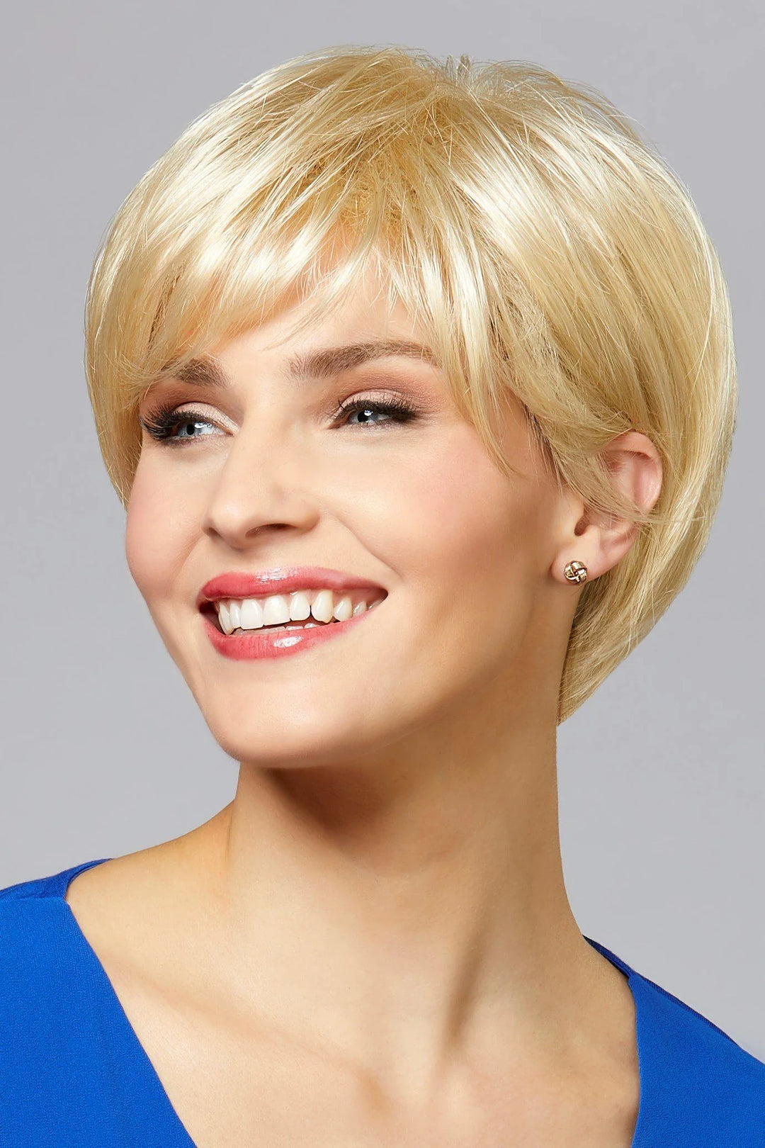 A smiling woman with a short blonde pixie wig, wearing a bright blue top, photographed against a solid gray studio background.