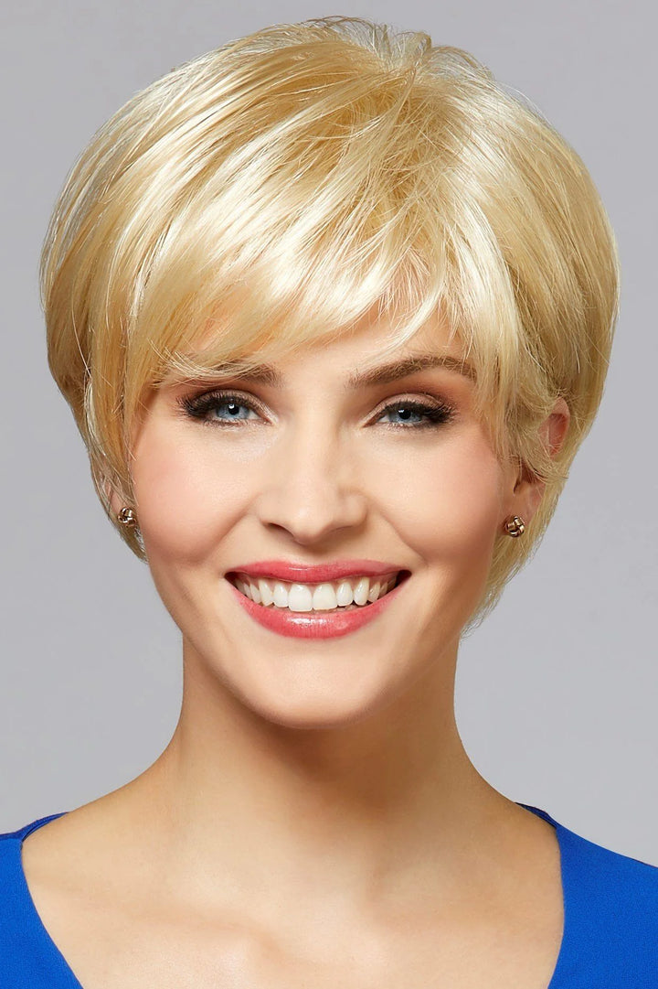 A smiling blonde woman with a short layered pixie wig, shown from the chest up against a light gray studio background.