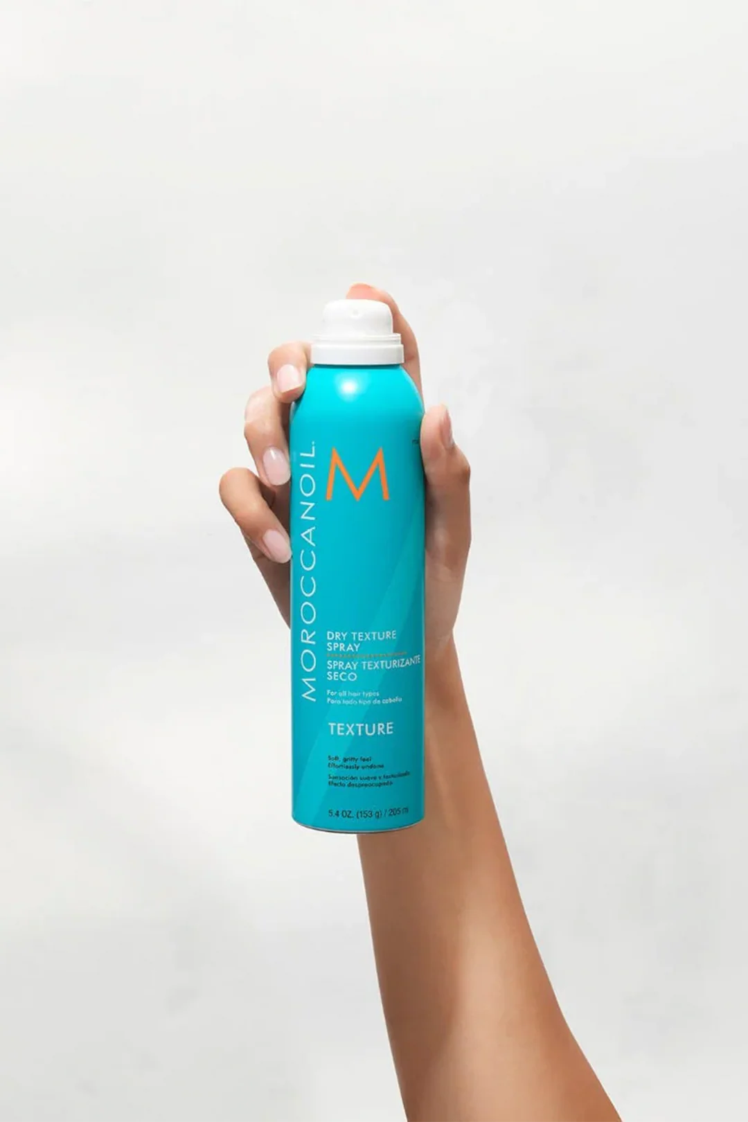 Hand holding a blue bottle of Moroccanoil hair product against a light gray background