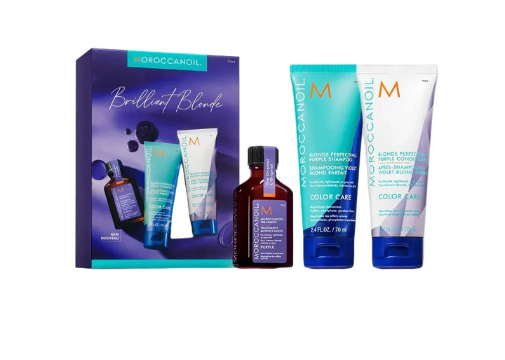 GROCCANOIL Brilliant Blonde hair color gift set featuring a purple box, a small amber bottle, a turquoise tube, and a white tube with blue caps, arranged on a light stone surface against a warm off-white background.