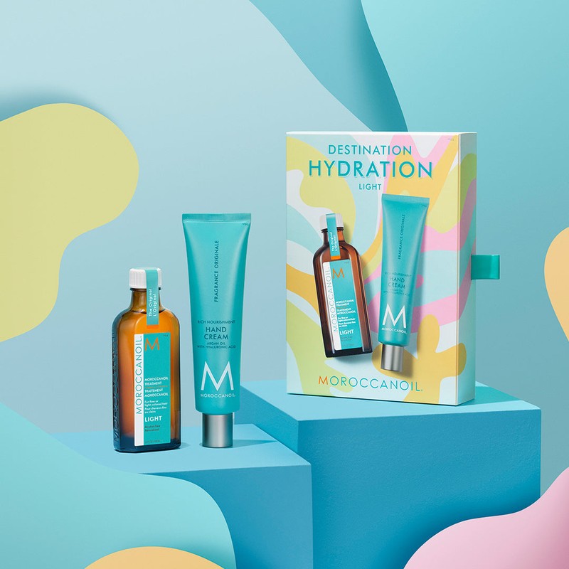 Moroccanoil Destination Hydration Light skincare set including a brown amber bottle, a turquoise hand cream tube, and its colorful packaging box, displayed on blue geometric pedestals.